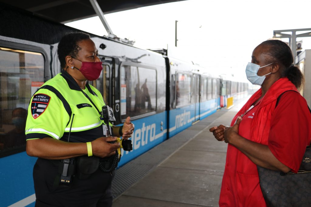 Security personnel talking to a passenger on a MetroLink platform with a blue train passing behind it.